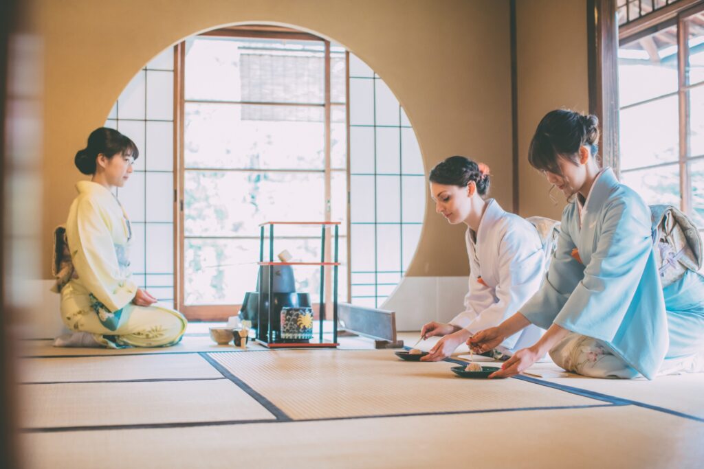 Three women in traditional Japanese kimonos participating in a tea ceremony inside a tatami room. The room is elegantly decorated with shoji screens and natural light filtering through the windows, highlighting the serene ambiance of the ceremony.