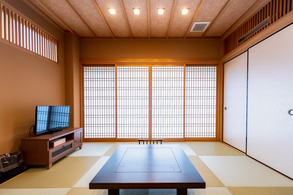 A modern Japanese tatami room with a minimalist design. The room includes a low wooden table, a flat-screen TV on a stand, and shoji screens covering the windows, providing a serene and well-lit environment.