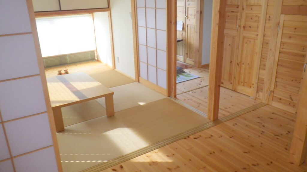 A cozy Japanese room featuring a mix of wooden and tatami flooring. The room is illuminated by natural light from a window, with a low wooden table and sliding shoji doors creating a warm and inviting atmosphere.