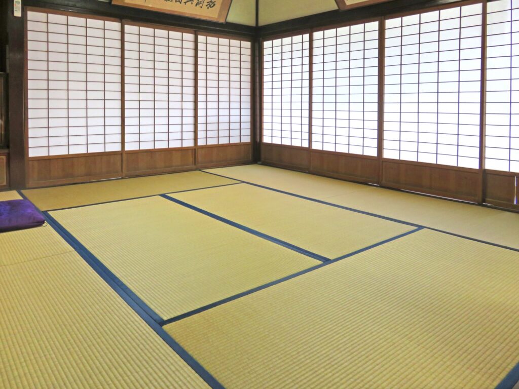 A traditional Japanese tatami room with large shoji screens covering the walls. The room features neatly arranged tatami mats and a tranquil, minimalist aesthetic typical of traditional Japanese interiors.