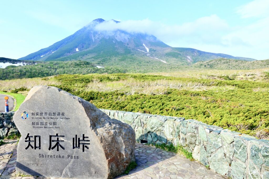 A scenic view of Shiretoko Pass in Shiretoko National Park, with a large stone marker in the foreground displaying the location's name in both Japanese and English, and lush green vegetation and a mountainous landscape in the background.