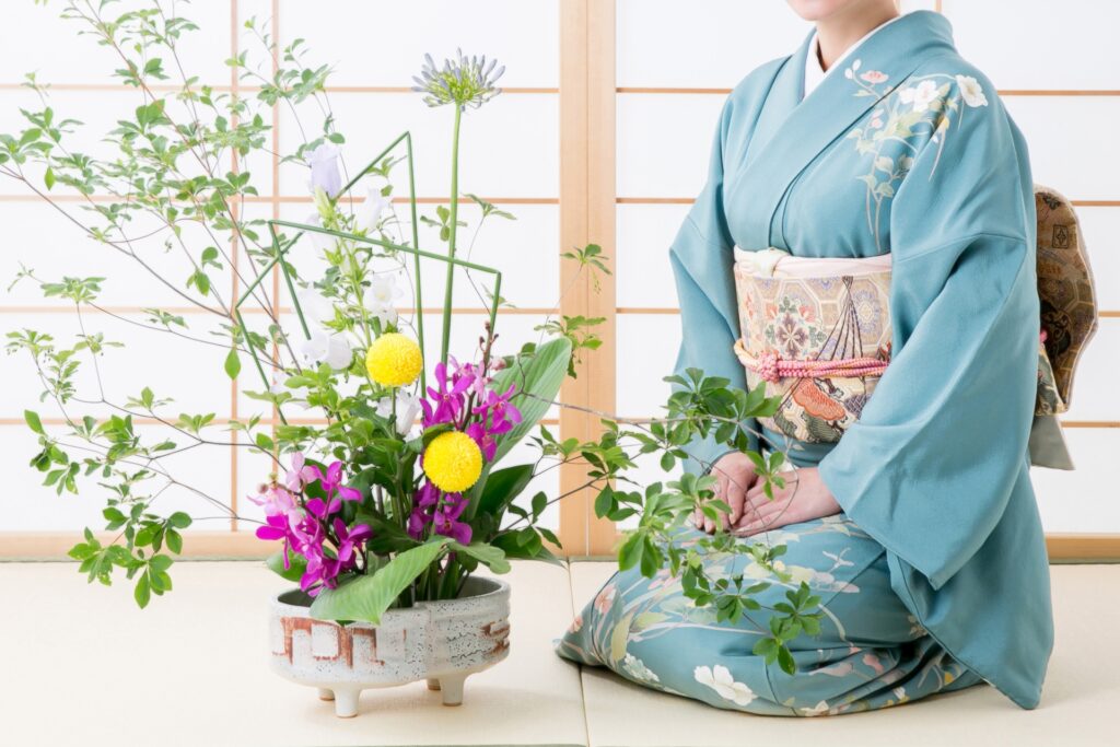 A person arranging a colorful floral bouquet, including sunflowers and various green foliage, in a basket. The scene is vibrant and lively, with other floral arrangements and greenery visible in the background.