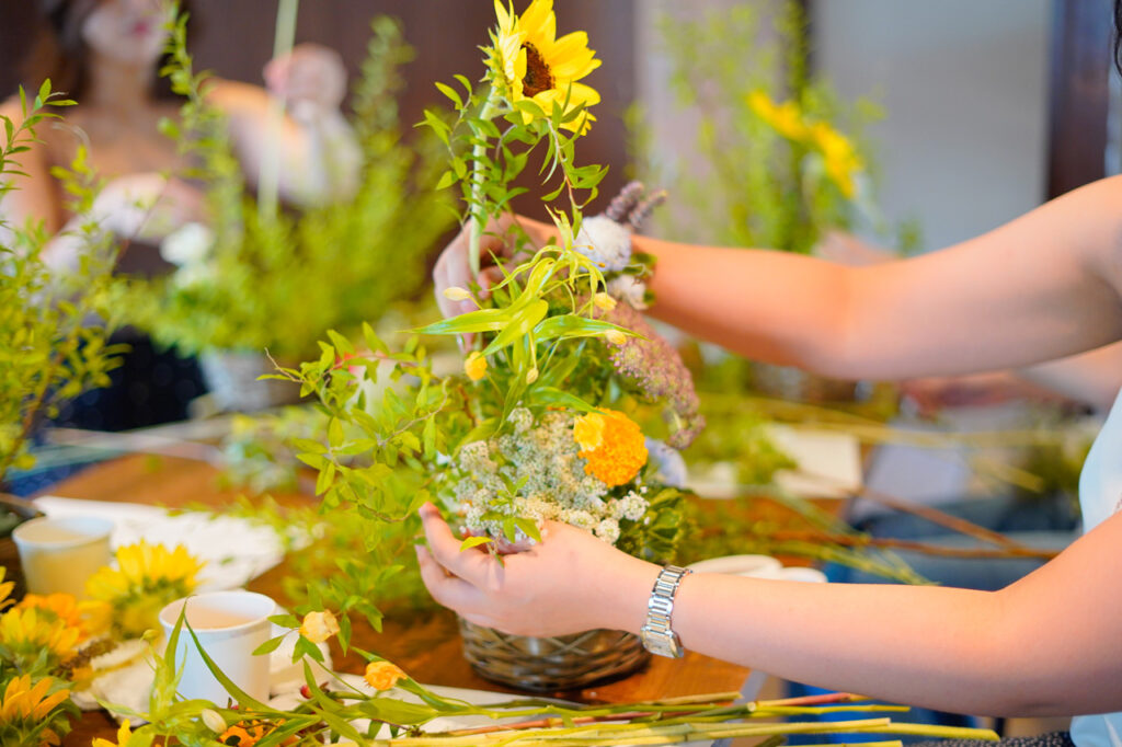 A person arranging a colorful floral bouquet, including sunflowers and various green foliage, in a basket. The scene is vibrant and lively, with other floral arrangements and greenery visible in the background.