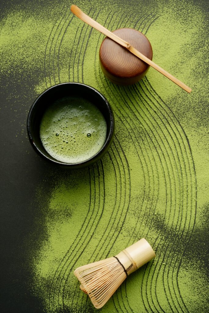 A bowl of matcha tea with a chasen (bamboo whisk) on a dark surface, surrounded by artistic swirls of matcha powder, creating a visually appealing and serene composition.