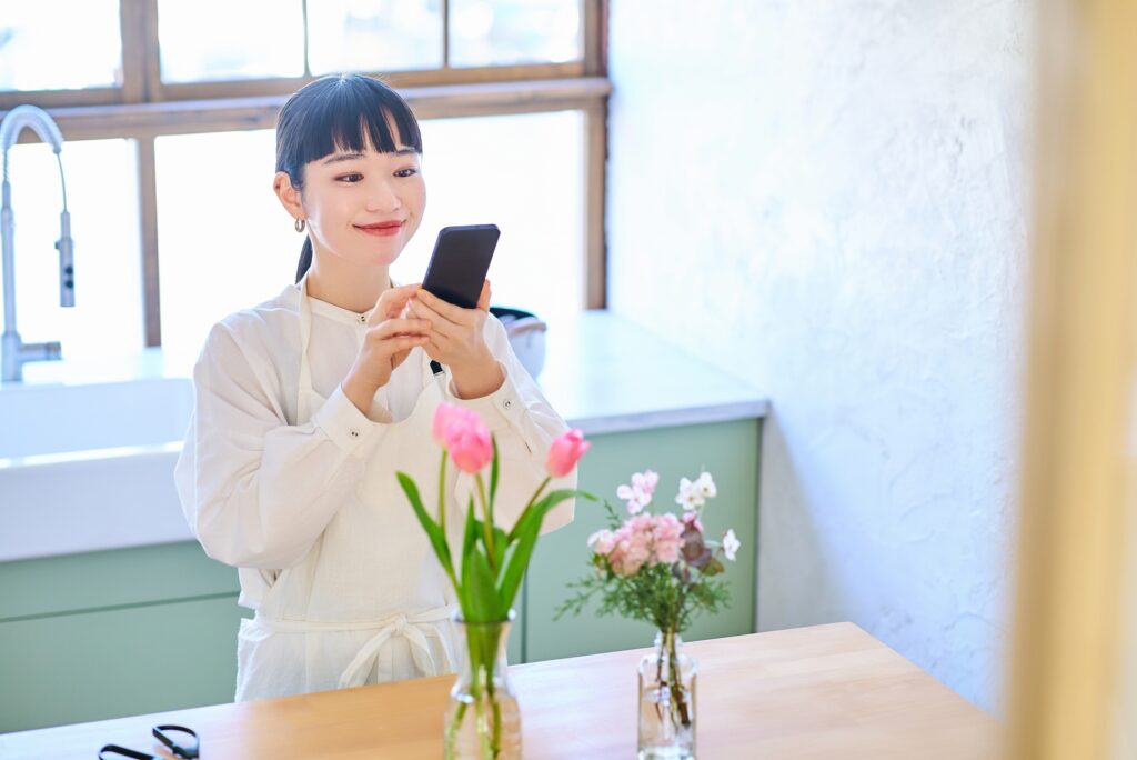 A young woman wearing a white apron standing in a bright kitchen, looking at her smartphone. She is smiling and appears to be taking a photo or checking her messages, with a vase of pink and white flowers on the table in front of her.