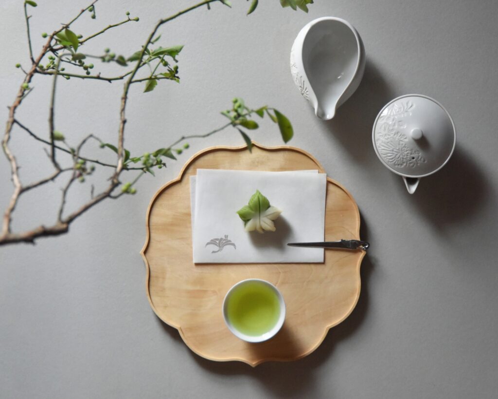 A beautifully arranged tea setup featuring a cup of green tea, a piece of wagashi (Japanese confectionery) on a wooden tray, and a branch with leaves, showcasing the elegance and simplicity of Japanese tea culture.