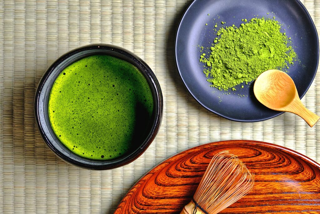 A top-down view of a bowl of matcha tea on a tatami mat, accompanied by a plate of matcha powder with a wooden scoop and a chasen on a wooden tray, highlighting the traditional elements of matcha preparation.